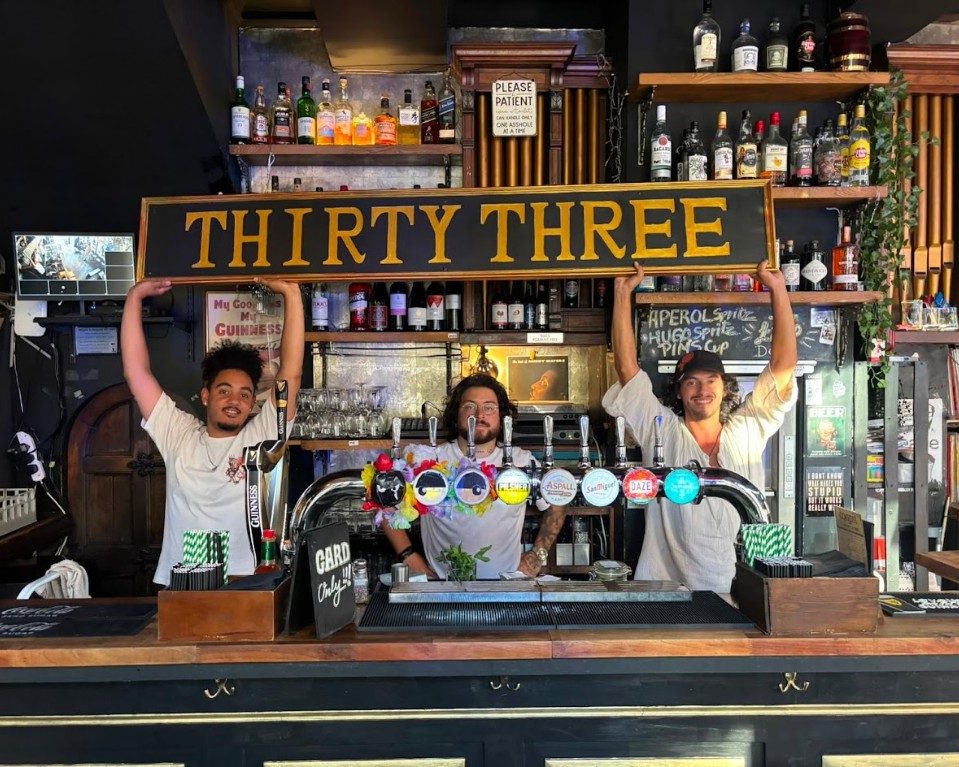 Three of the bar team holding the Thirty Three sign above their heads behind the main bar